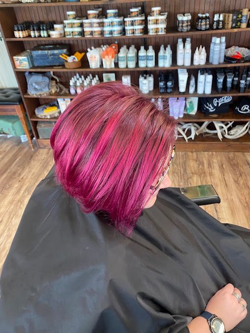 Client with bright pink hair in a salon chair, seen from behind, with shelves of hair products in the background