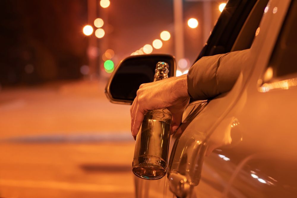 A person is leaning out of a car window holding a bottle of beer.