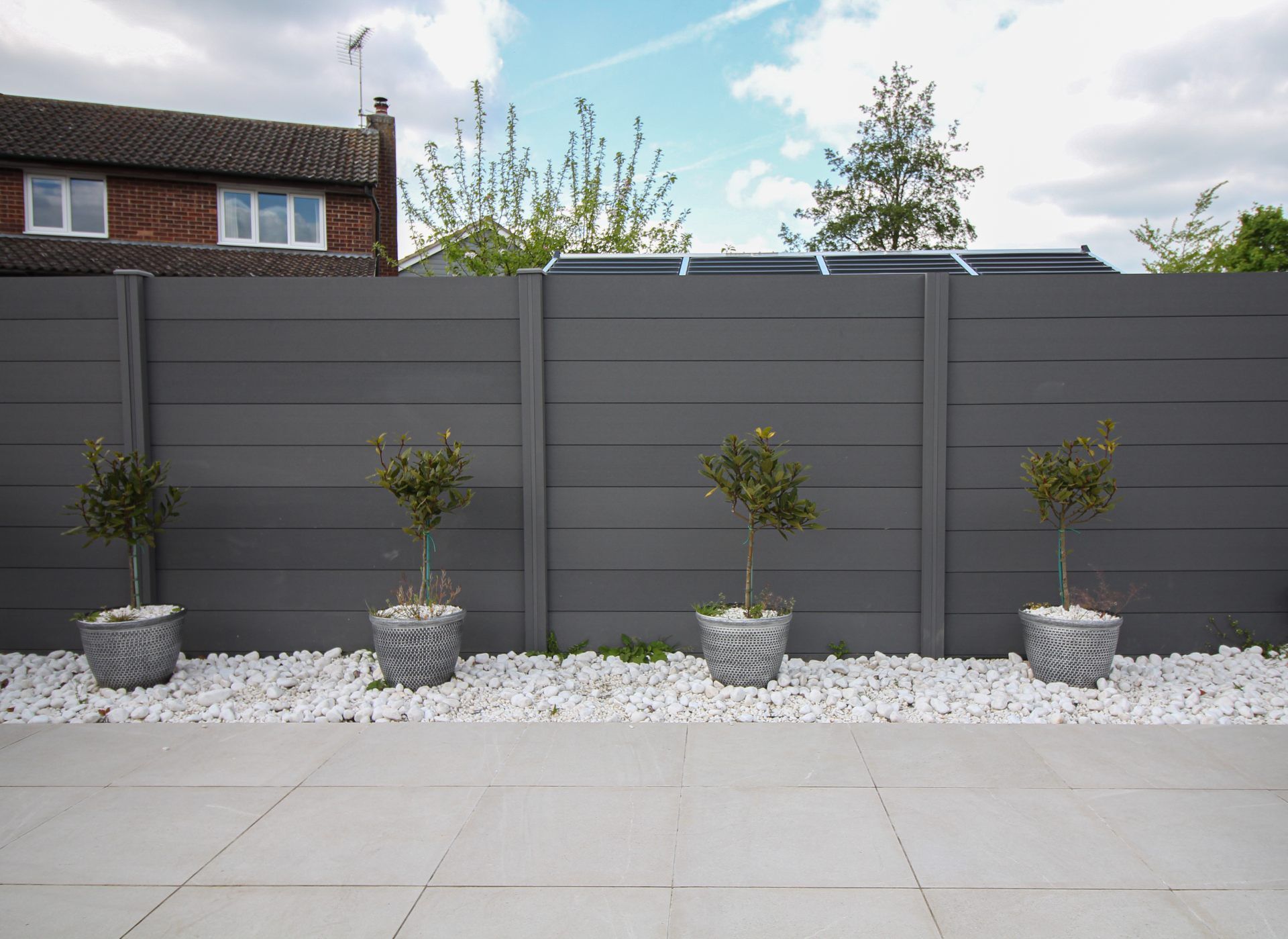 Modern gray fence with four potted plants on white gravel beside a tiled patio