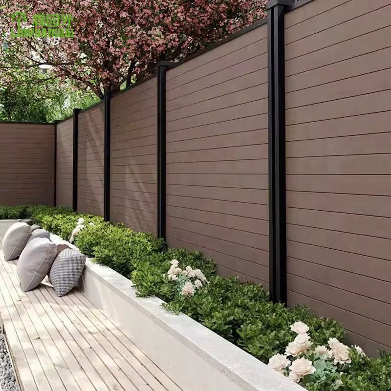 Modern backyard patio with tan privacy fence, white planter beds, and shrubs with cushions along a wood deck