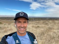 A man wearing a hat and a backpack is taking a selfie on top of a hill.