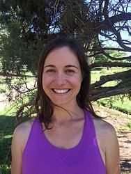 A woman in a purple tank top is smiling in front of a tree.