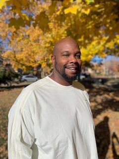 A man in a white shirt is standing in front of a tree with yellow leaves.