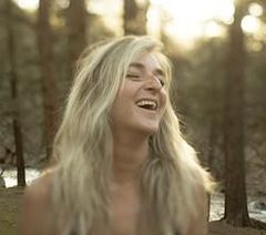 A woman is laughing in the woods with her eyes closed.