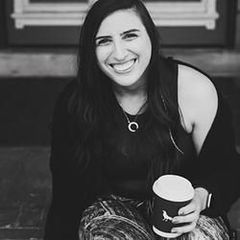A woman is sitting on the ground holding a cup of coffee and smiling.