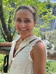 A woman in a white dress is smiling in front of a waterfall.