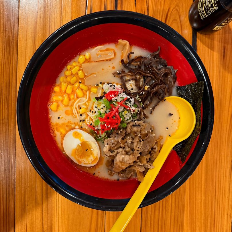 Red ramen bowl with corn, egg, mushrooms, minced meat, and a yellow spoon on a wooden table