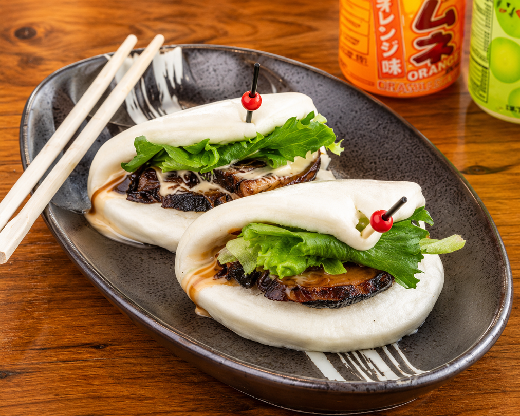 Two steamed buns filled with meat and greens on a dark plate, with chopsticks and an orange drink nearby