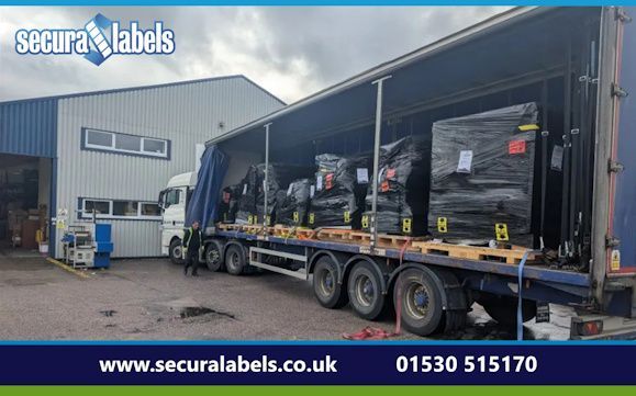 Shiny new kit arriving A HGV is parked up outside a light grey clad factory unit with its side curtains pulled open revealing stacks of palletised goods wrapped in black plastic. A person in a high-visibility vest is walking along the side of the trailer. The Secura Labels logo is overlaid in the top left hand corner of the image against a grey cloudy sky. At the bottom of the image is a dark blue band containing white text that says ‘www.securalabels.co.uk’ and a phone number of ‘01530 515170’.