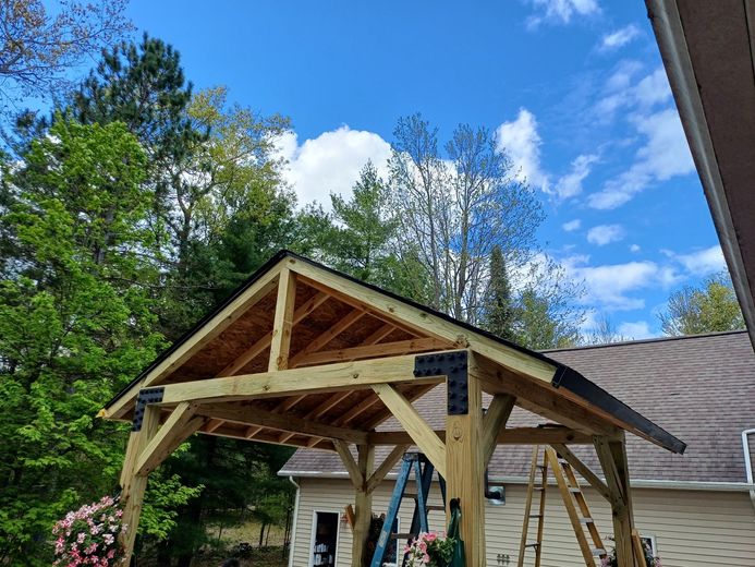 Wooden gazebo under construction in a backyard, with ladder and roof framing against a blue sky.
