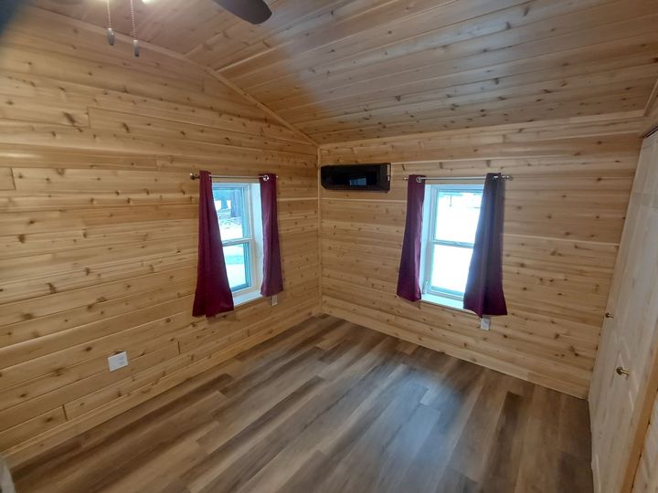 Small wooden room with two windows, red curtains, and a wood floor and ceiling.