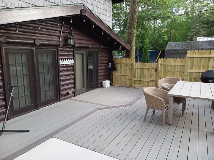 Wooden deck patio beside a rustic cabin, with wicker chairs, a table, and a fenced yard.