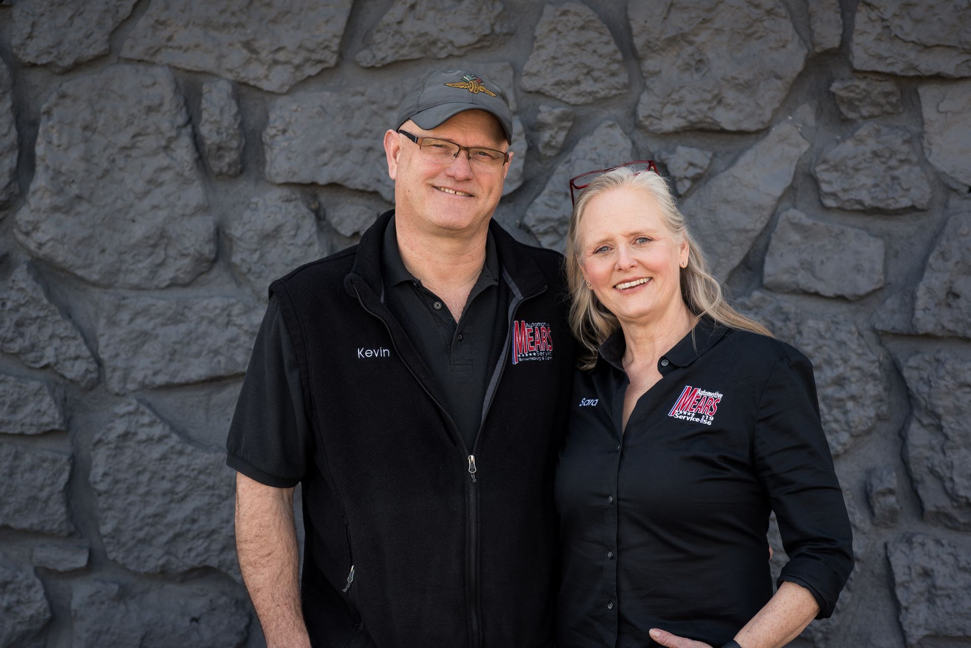 Two smiling people in black shirts standing in front of a dark stone wall | Mears Automotive