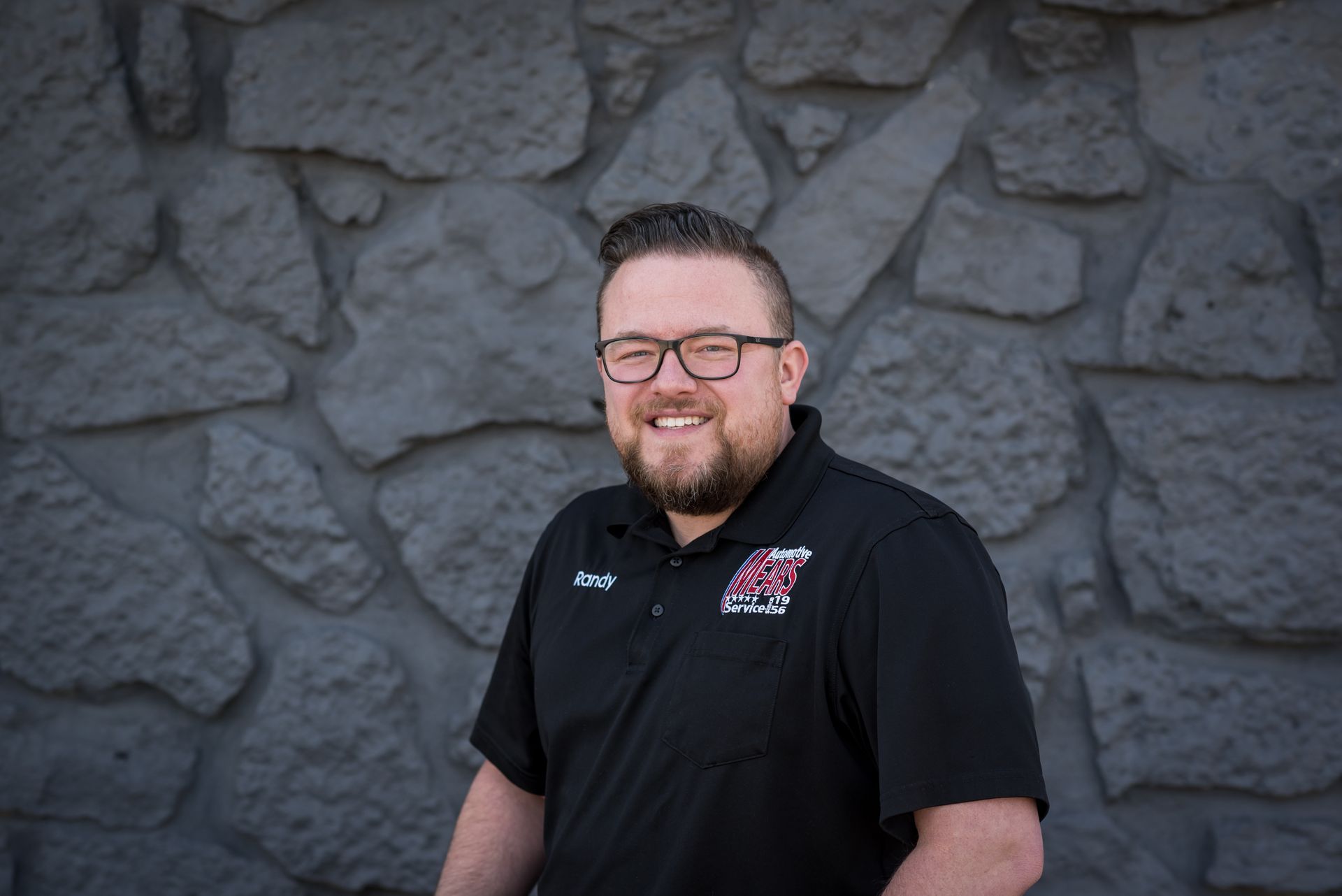 Smiling man in a black collared shirt standing before a gray stone wall | Mears Automotive
