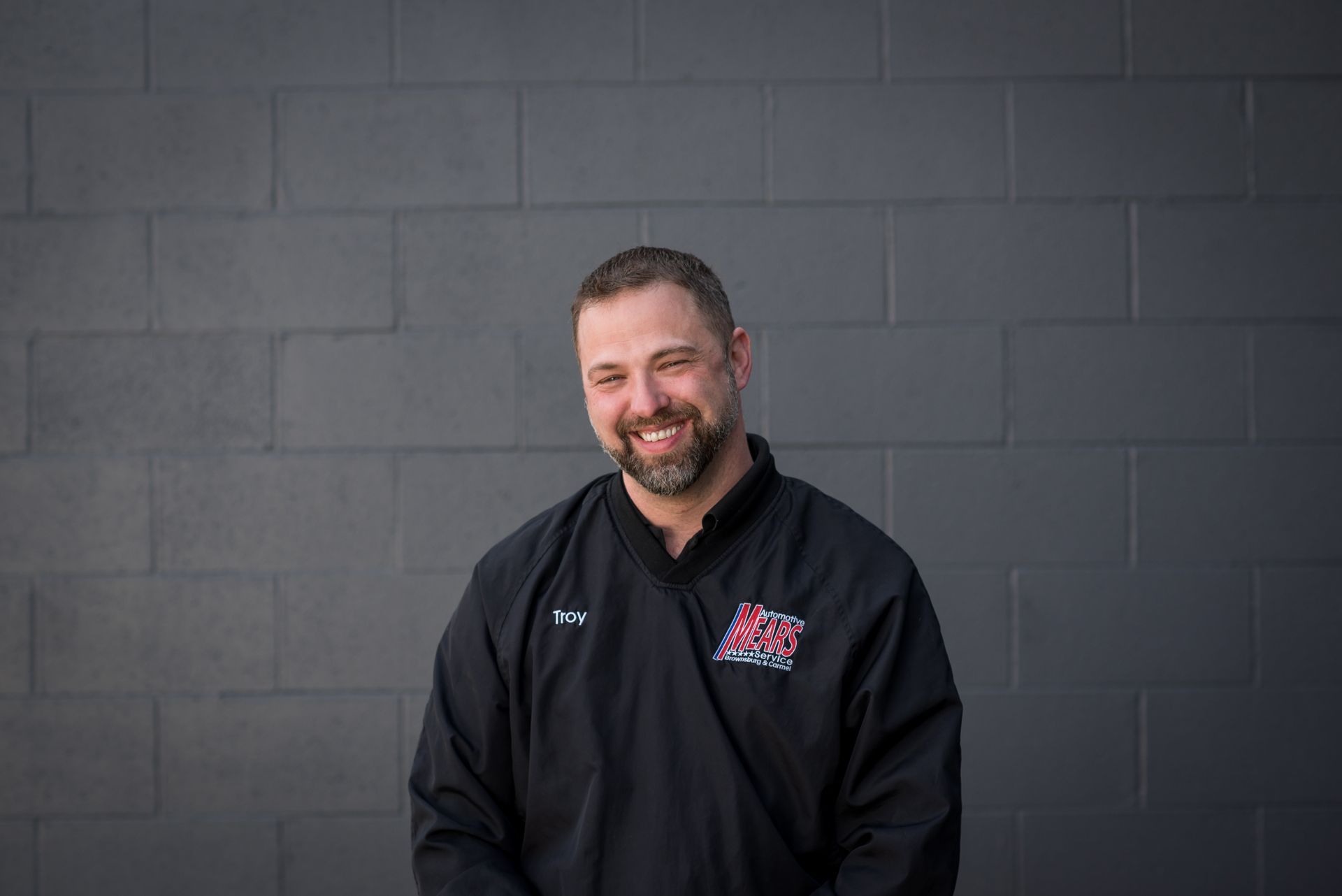 Smiling man in a black jacket standing against a dark gray brick wall. | Mears Automotive