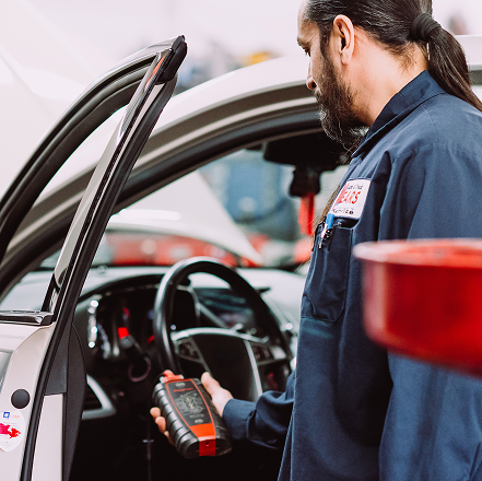 Mechanic holding a diagnostic scanner beside an open car door in an auto repair shop | Mears Automotive