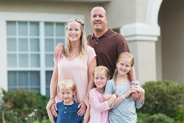 Family of four standing outside a home, smiling and hugging, with a white house and greenery behind them