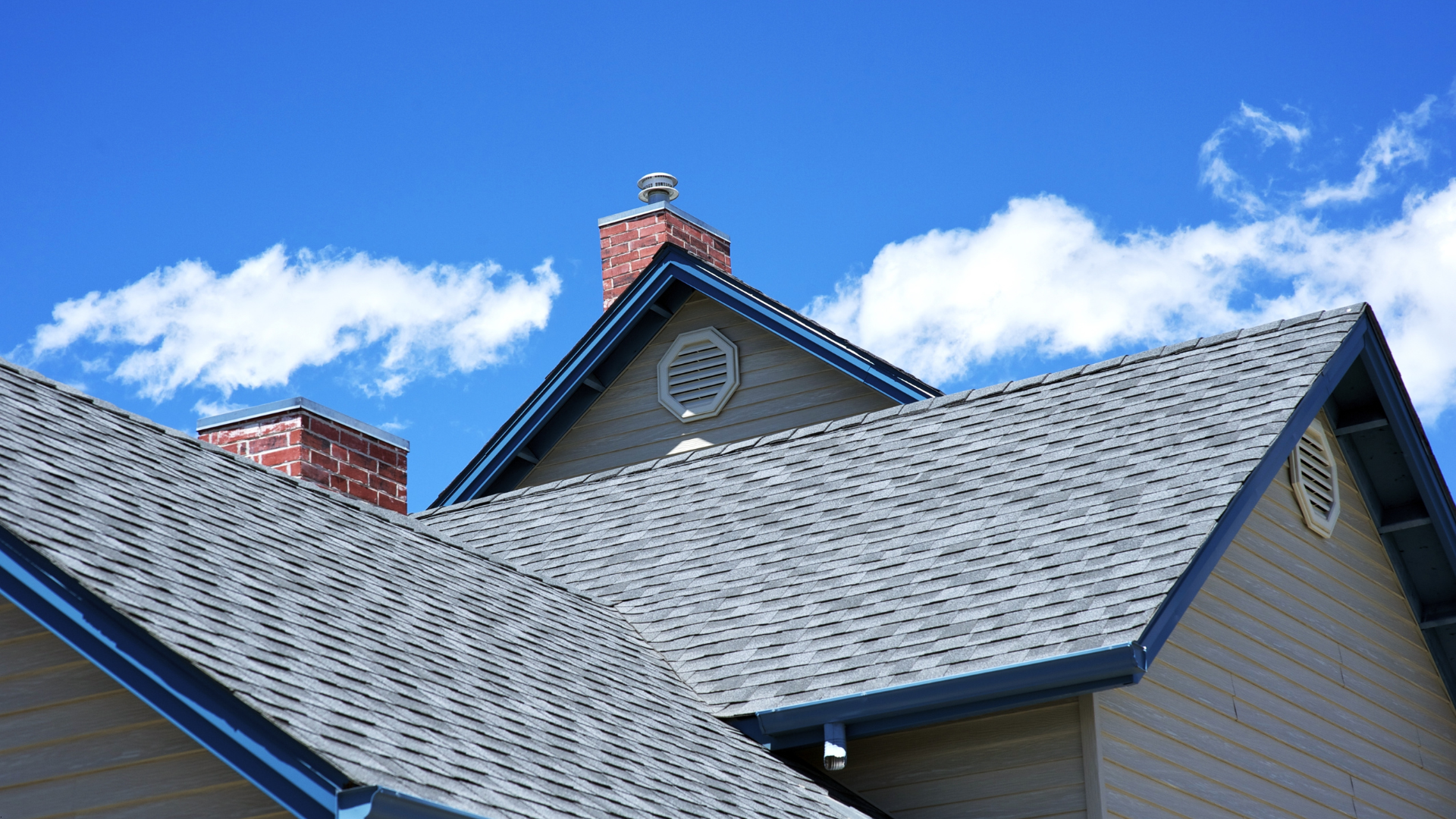 Gray shingled rooftops and brick chimneys against a bright blue sky with fluffy white clouds.