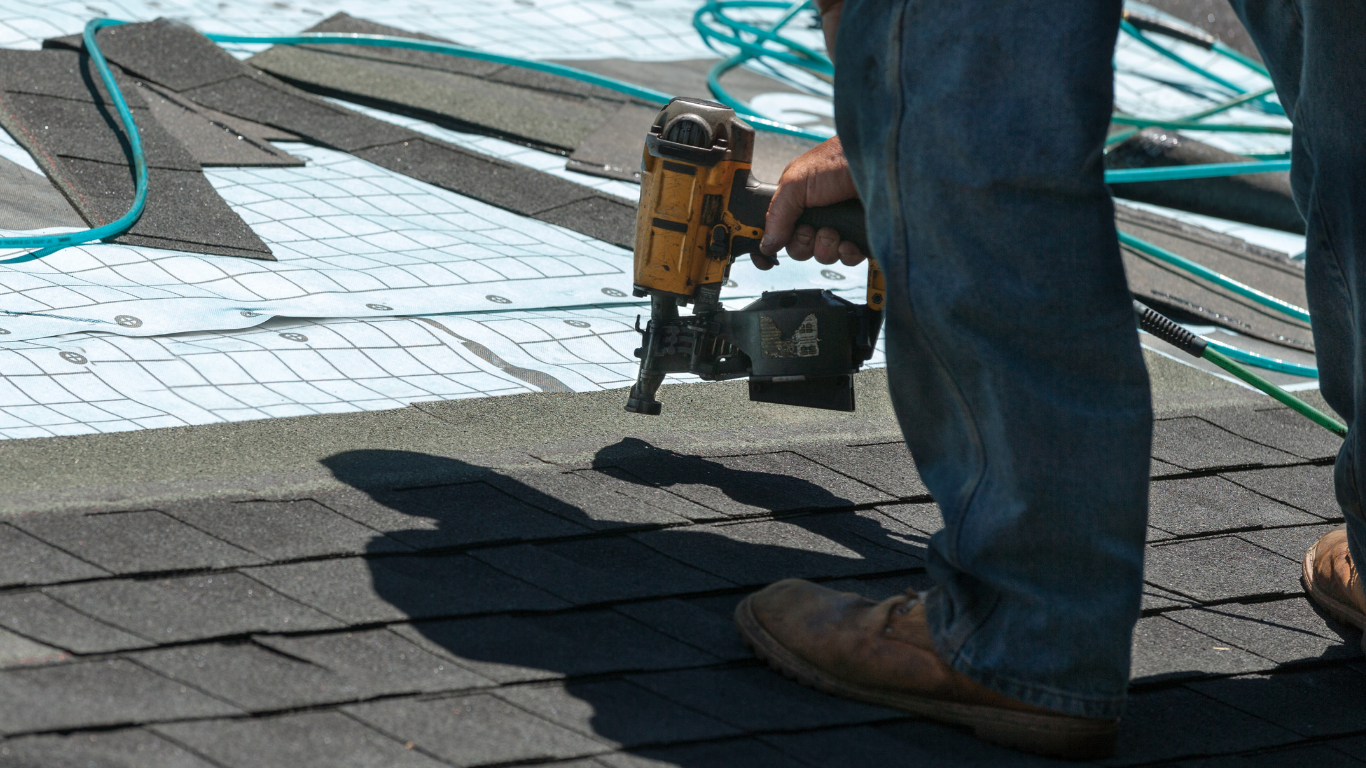 Roofer using a nail gun to install dark-colored shingles on a roof.