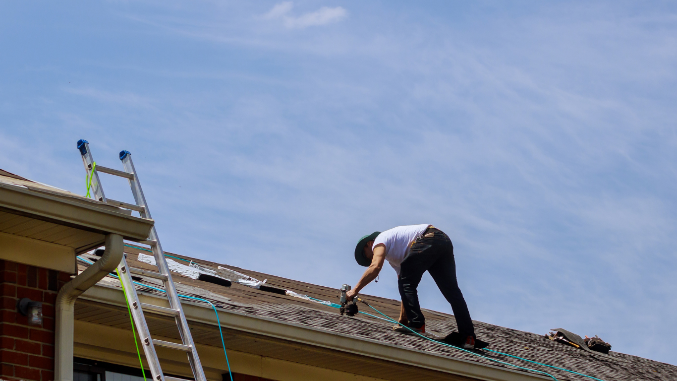 Person on a rooftop works on the shingles near a ladder. Sunny, daytime.