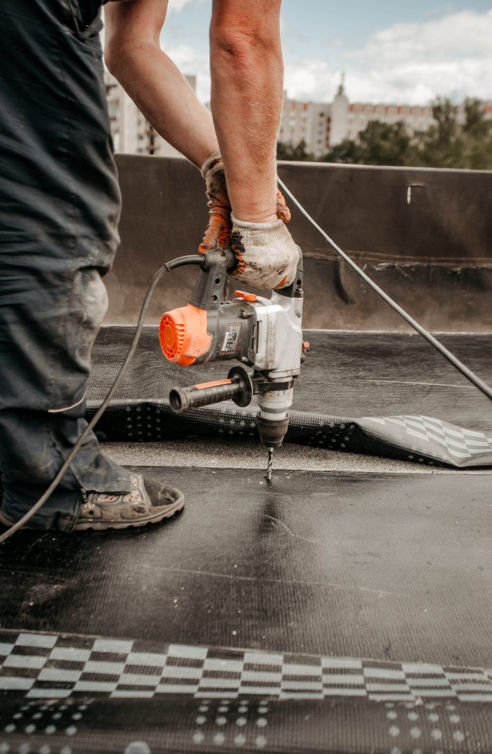 Person using a power drill on a dark, patterned surface outdoors.