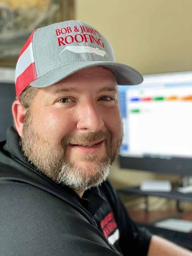 Smiling man in a red-and-gray cap and black shirt in an office, with a computer screen blurred behind him