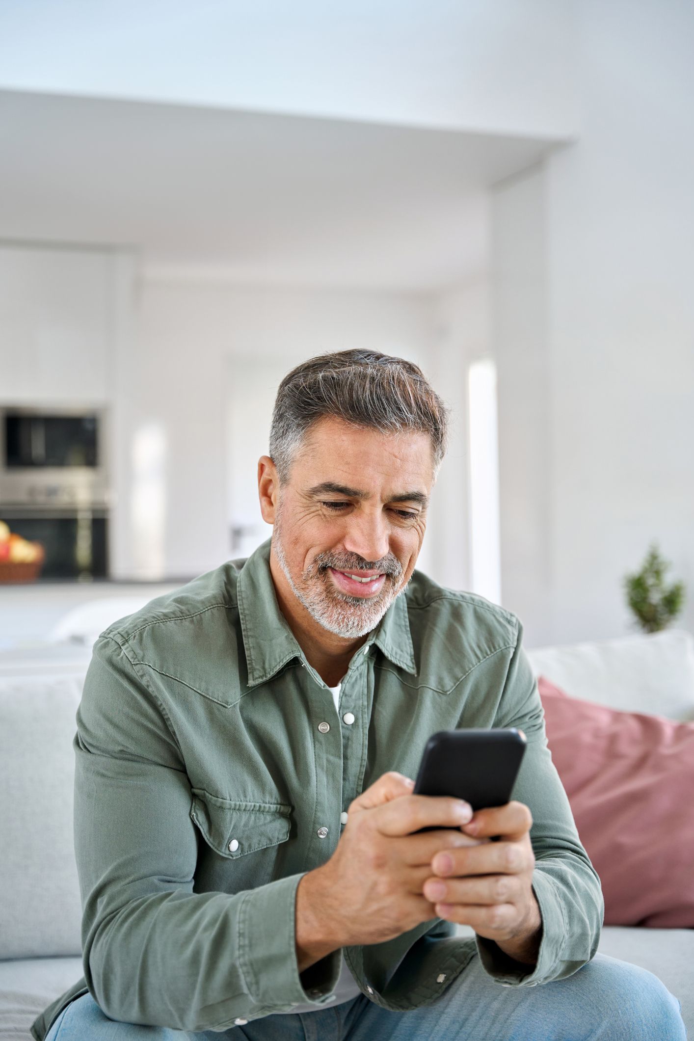 Man sitting on a couch, smiling while using a smartphone in a bright living room