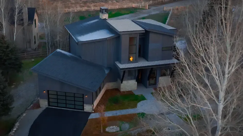Modern gray house with angled roof, garage, and walkway; trees and mountain in background.