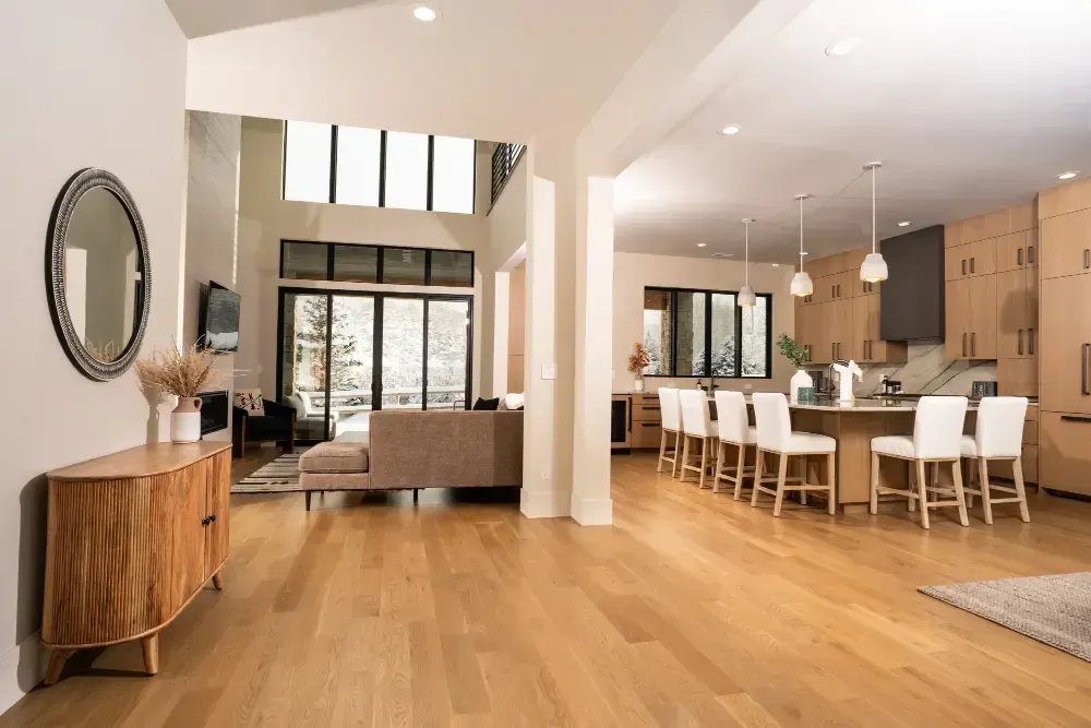 Open-concept living space with a kitchen, dining area, and living room. Light wood floors, white walls, and large windows.