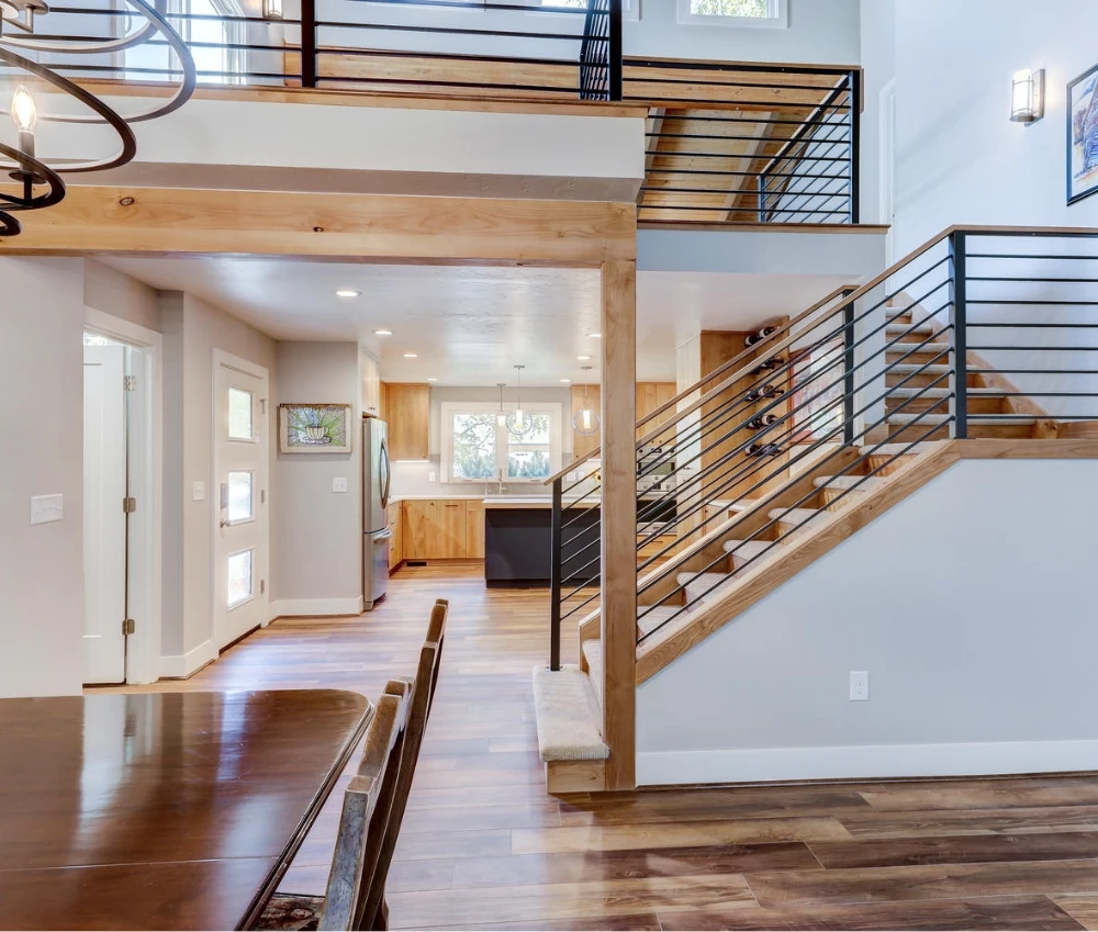 Interior view of a modern home with a staircase, kitchen, and dining area. Light wood and gray walls.