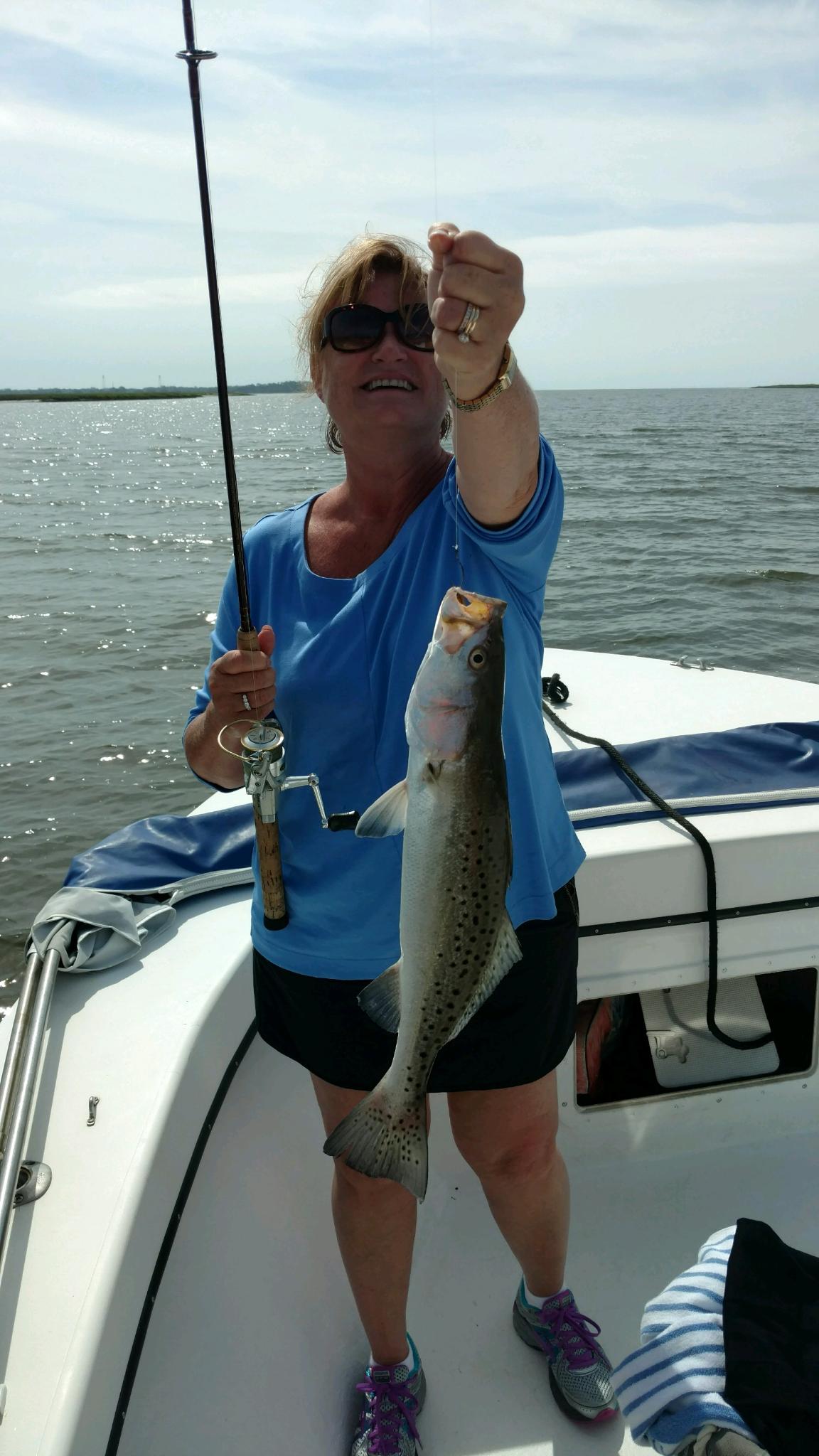 Woman on a boat holding up a speckled trout she caught; water and sky in the background.
