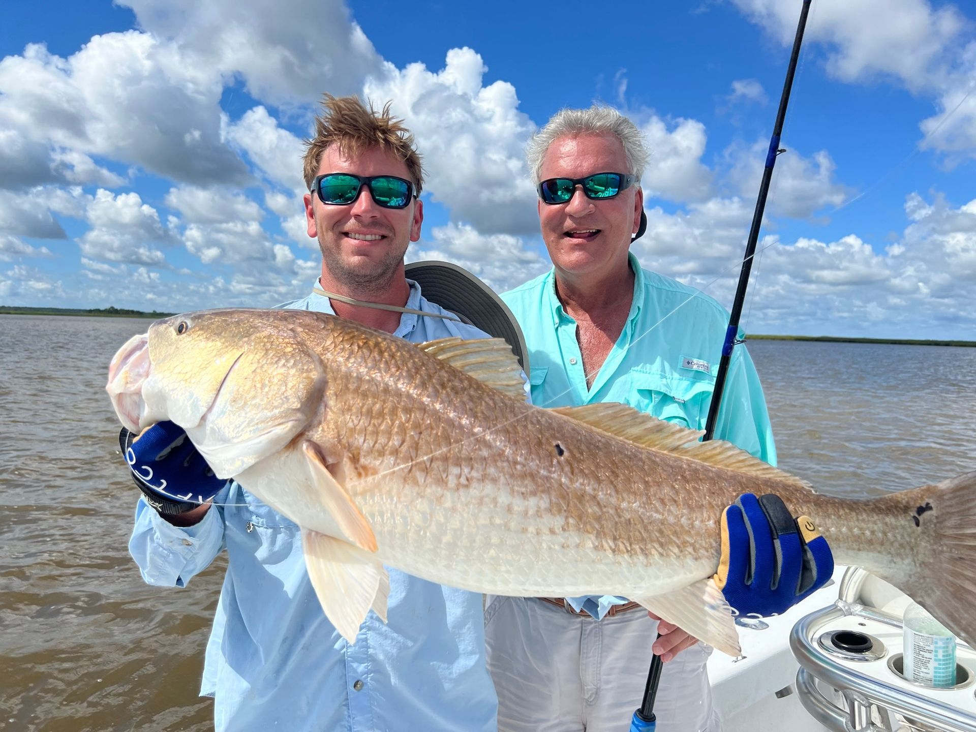 Two men holding a large redfish on a boat in a sunny bay.
