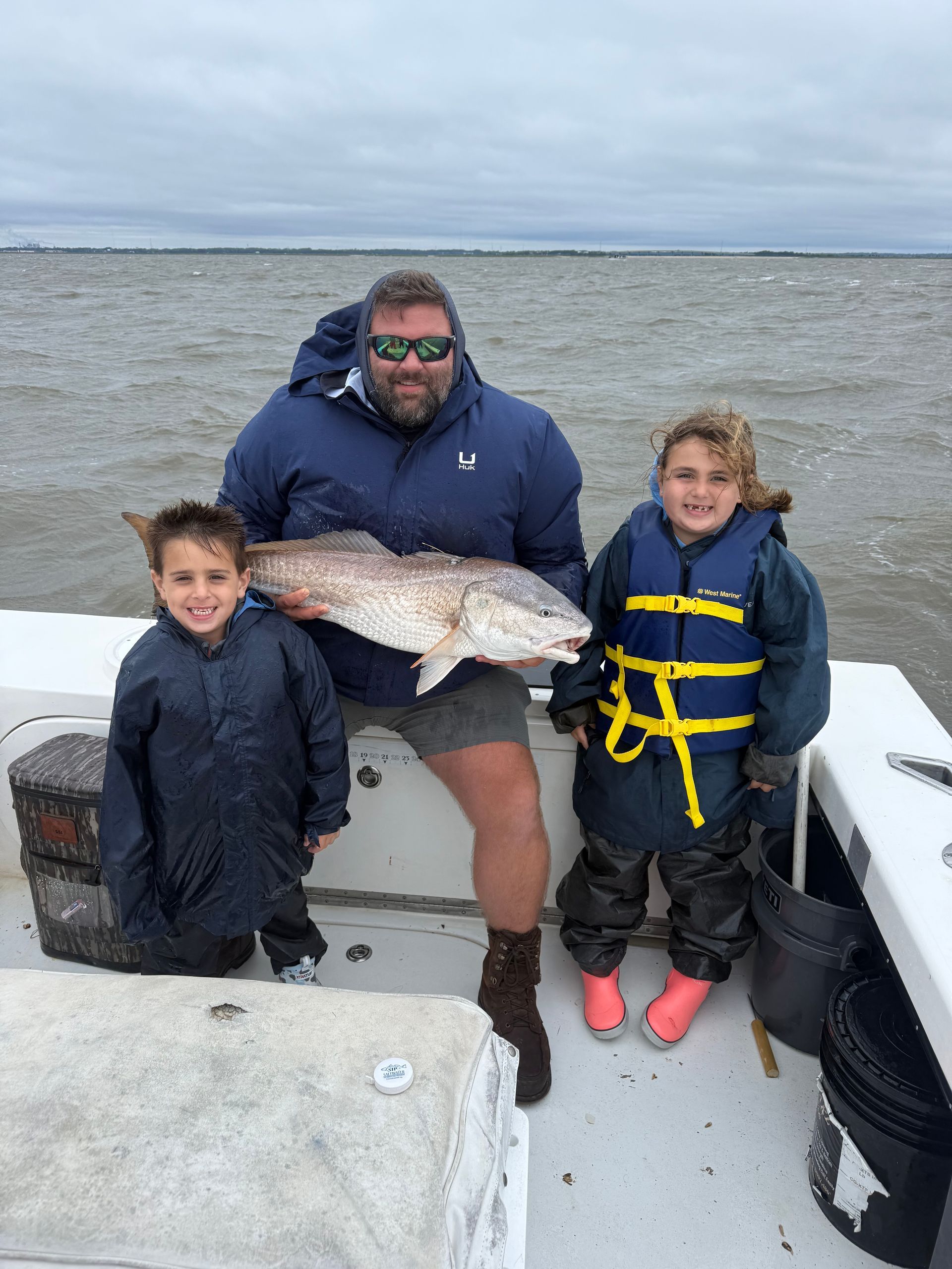 Man and two children on a boat holding a large redfish. Overcast day on the water.