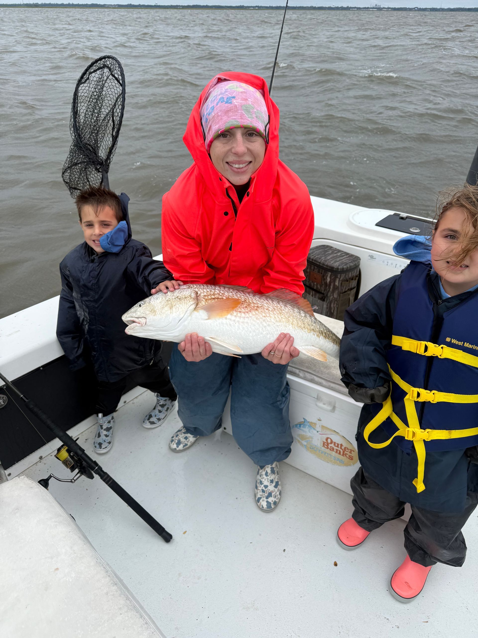 Woman on boat, holding large fish, with two children. Cloudy day, they're bundled up in jackets and boots.