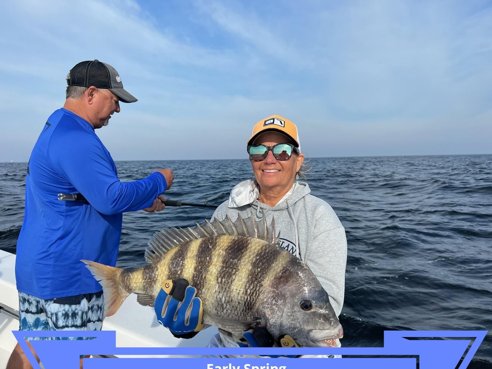 Woman on a boat smiles while holding a large fish, another person assists. Ocean in the background.