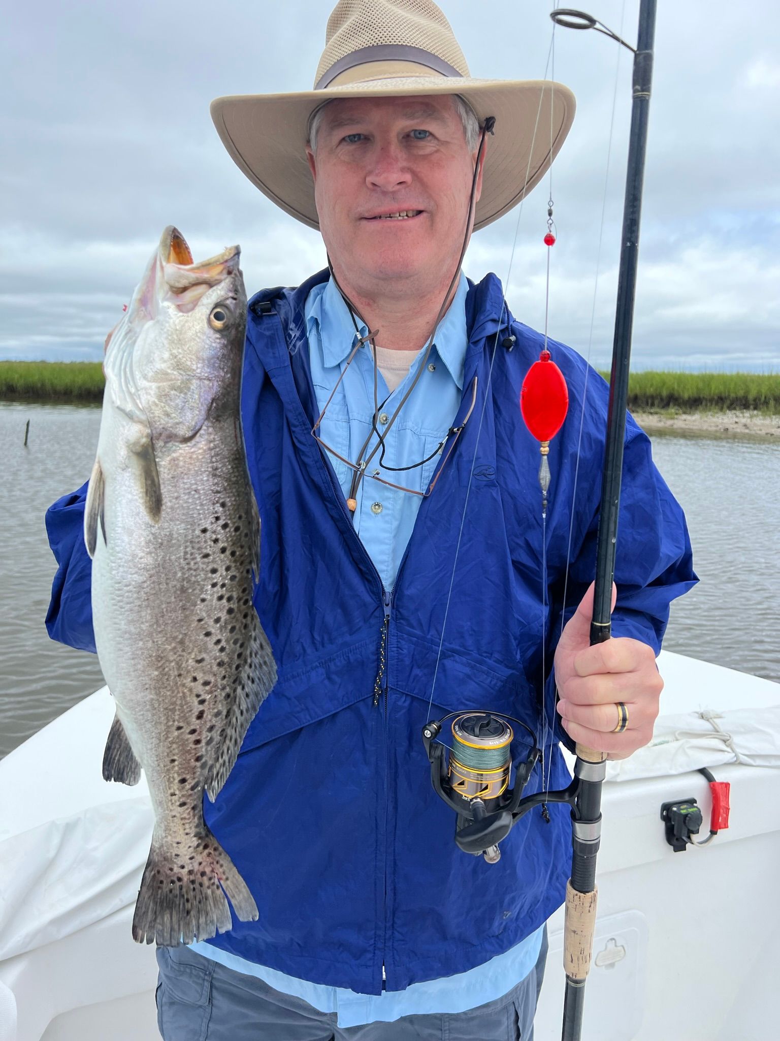 Man holding large fish, standing on a boat, wearing a hat and blue jacket, with a fishing rod.