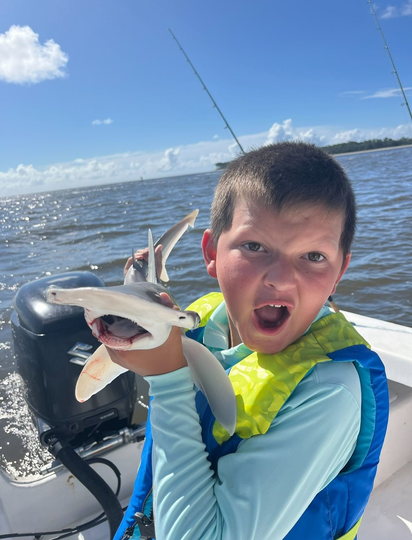 Boy on boat holding a small hammerhead shark, excited expression. Blue water, sunny sky.