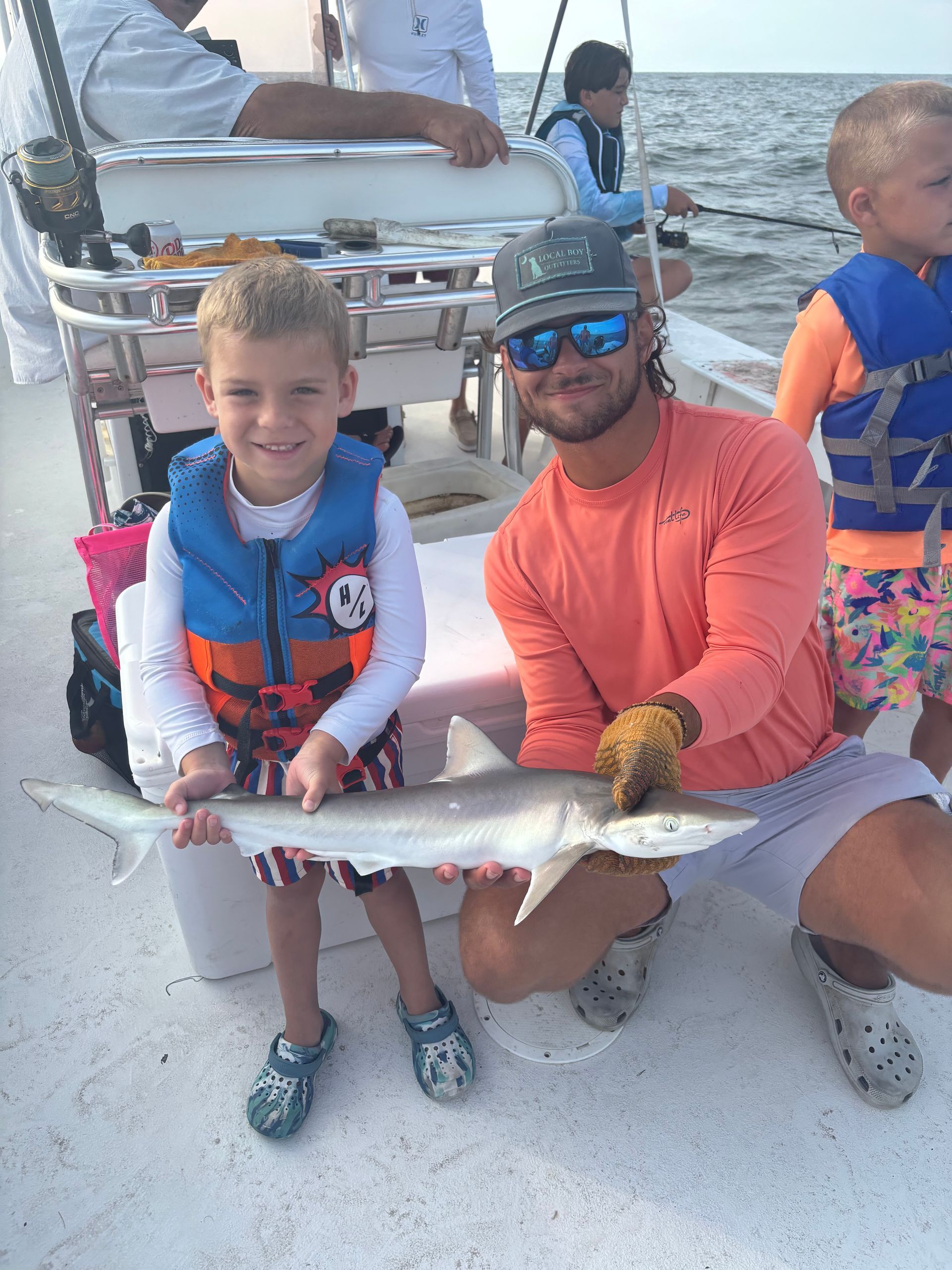 A man and a boy on a boat hold a small fish. The boy smiles, wearing a life vest.