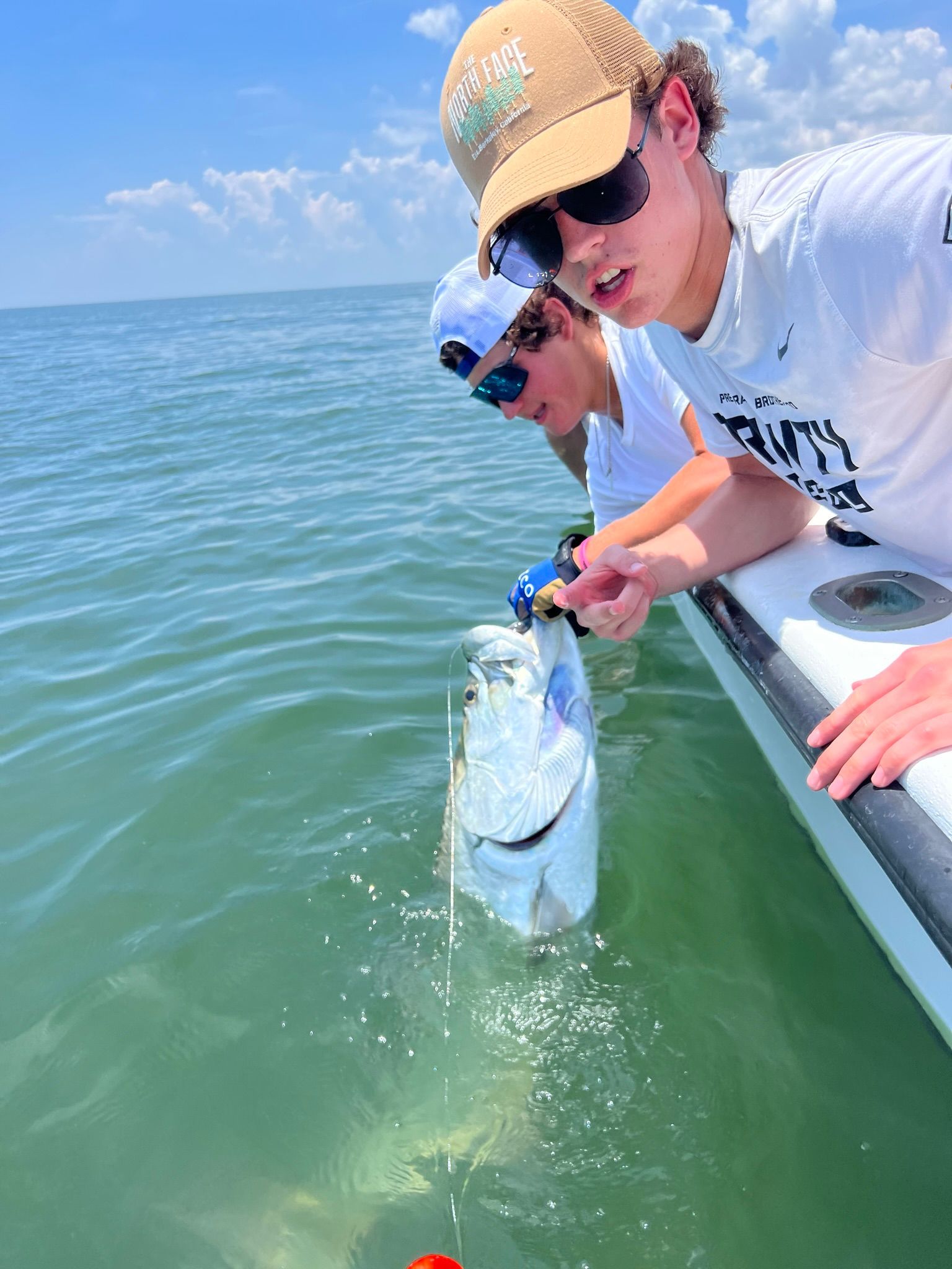 Two people on a boat holding up a silver fish in the water. One points at it. Blue sky.