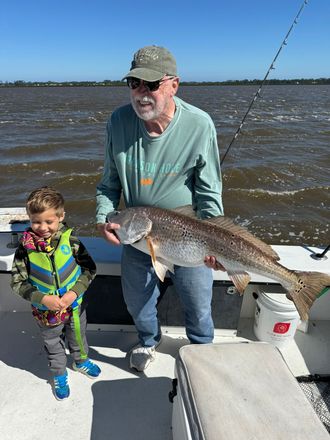 Man and child on a boat hold up a large fish; sunny day, water in the background.