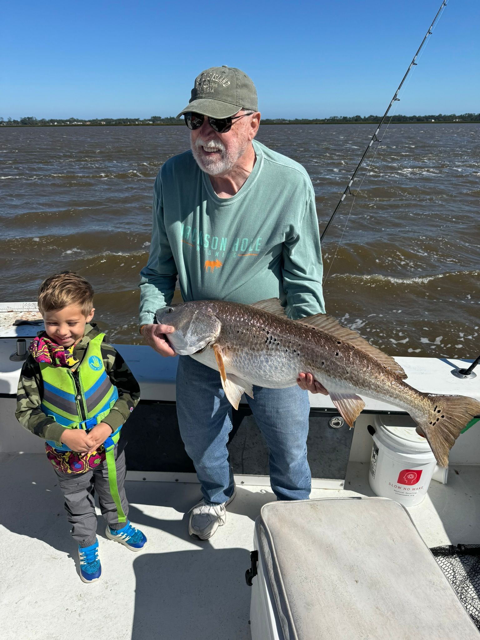 Man and child on a boat holding a large fish; blue water and sky background.