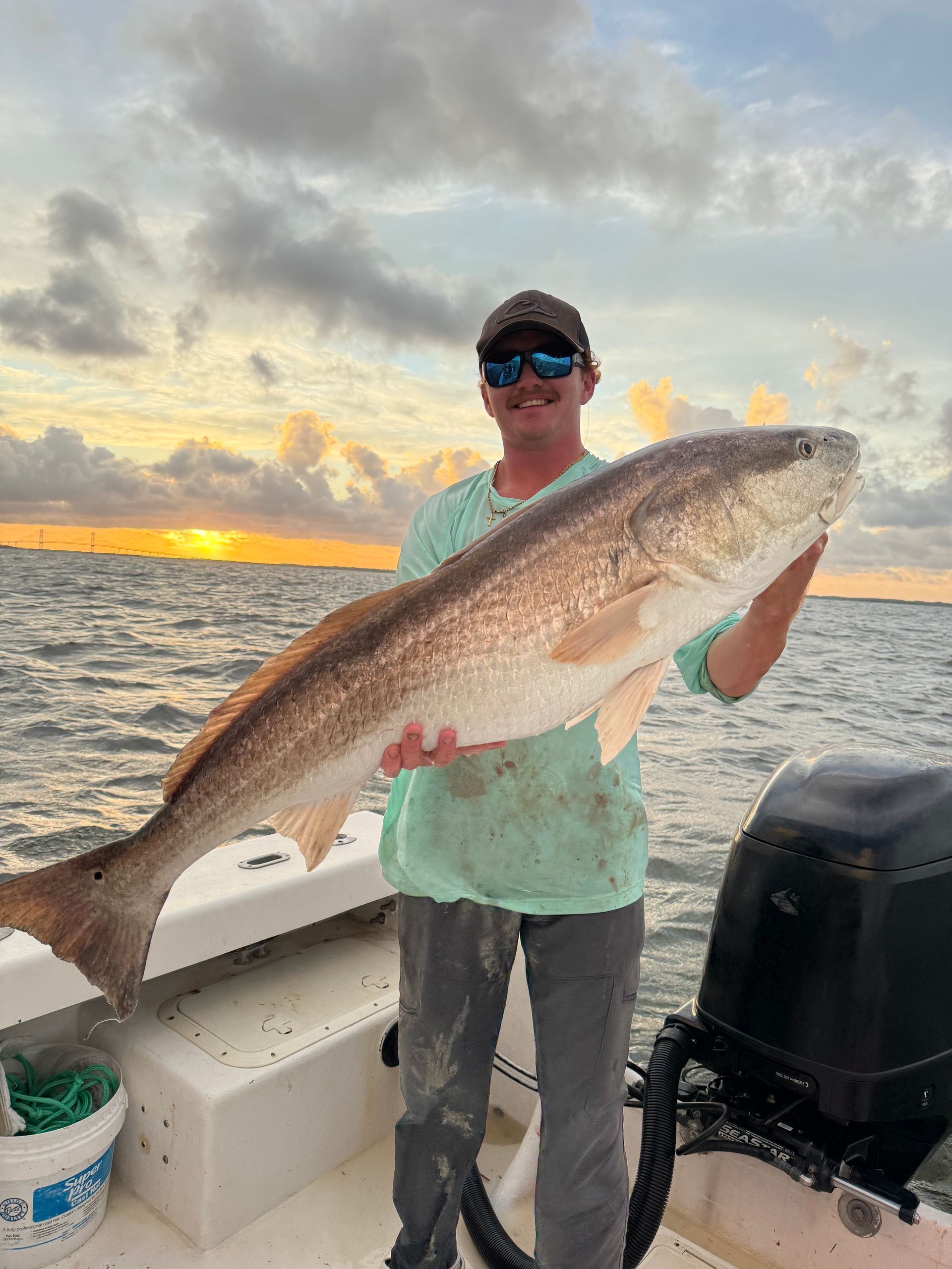 Man holding a large redfish on a boat, sunset in the background.