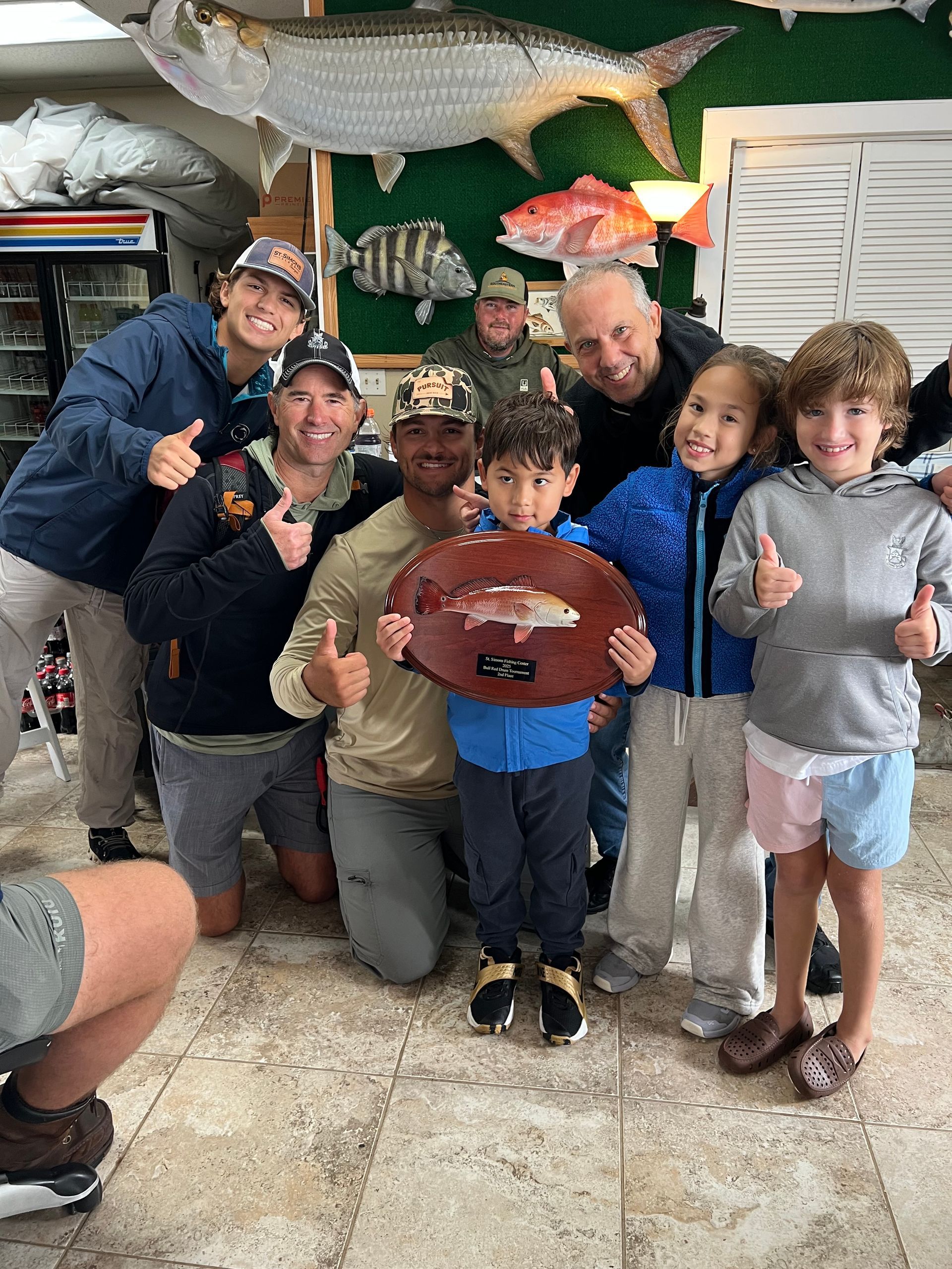Group of people posing with a fish trophy indoors, giving thumbs up.