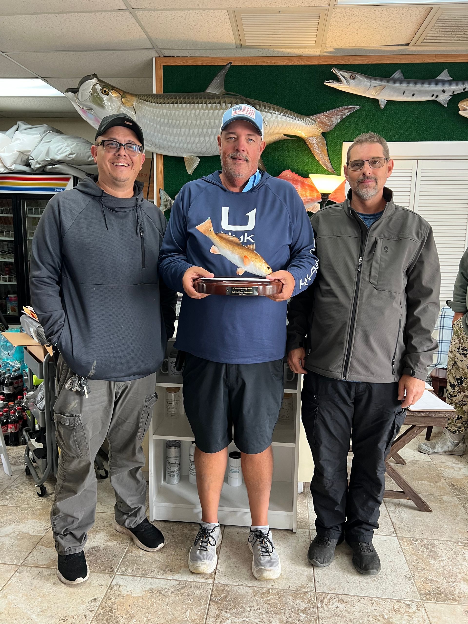 Three men with trophy, holding a fish model. Fishing store interior with mounted fish on the wall.