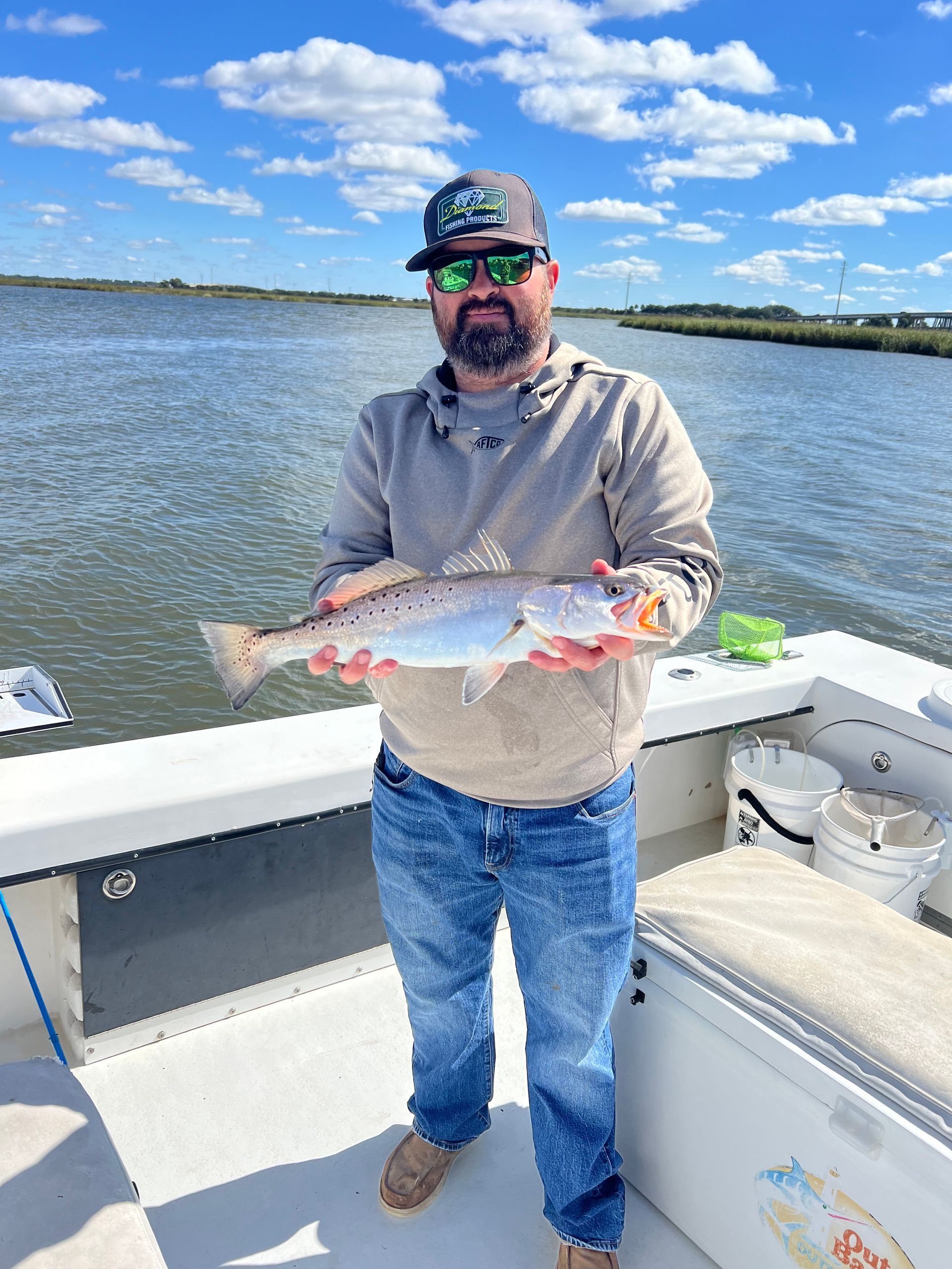 Man on a boat holds up a speckled trout he caught. He wears a hat, sunglasses, and jeans. Water and marsh in the background.