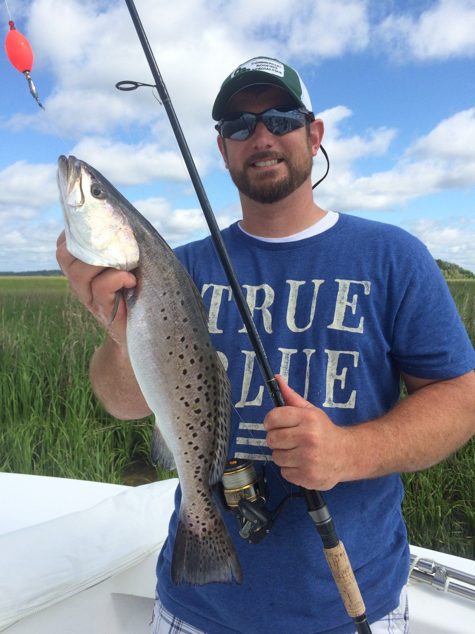 Man holding a speckled trout, smiling, wearing a hat and blue shirt. Fishing rod and bobber visible, marsh background.