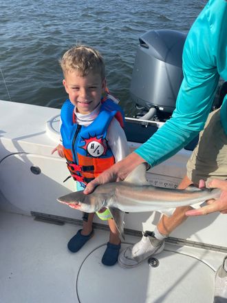 Boy in life vest holding a small shark on a boat, smiling. Water and outboard motor visible.