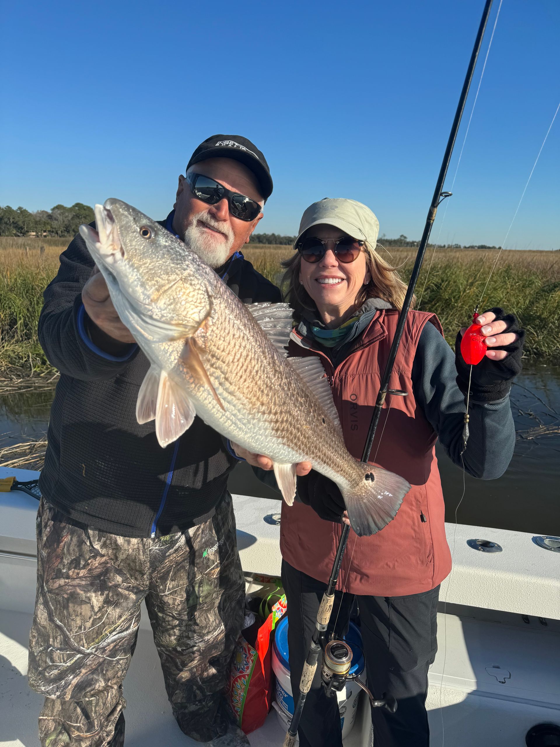 Man and woman on a boat hold up a large redfish with marsh grass in the background. Sunny day.