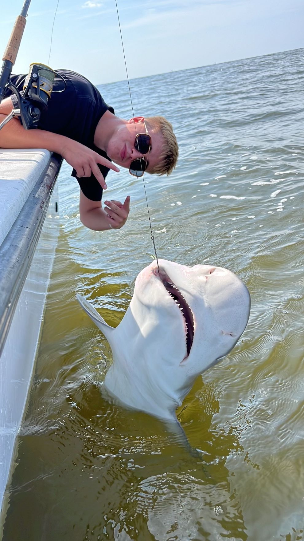 Young person on a boat catches a large, white fish. He holds the line while grinning, and gives a thumbs up.