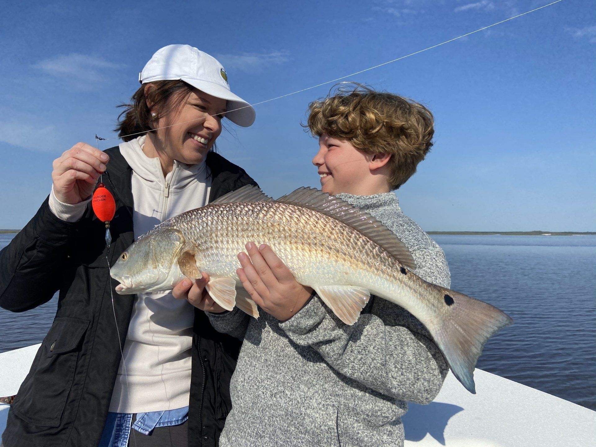 Caught with the Thunder Chicken in Louisiana-Bull red drum Woman and boy on a boat hold up a large redfish, smiling. Blue sky and water in the background.